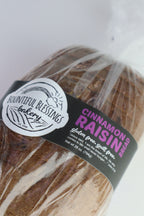 A loaf of Bountiful Blessings Bakery Cinnamon Raisin Bread wrapped in clear plastic with a black and white label featuring pink text.