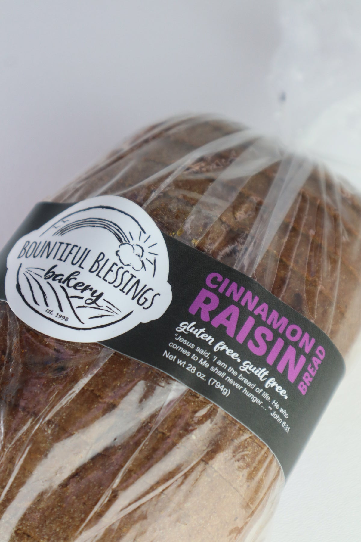 A loaf of Bountiful Blessings Bakery Cinnamon Raisin Bread wrapped in clear plastic with a black and white label featuring pink text.
