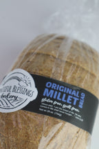 A close-up of sliced gluten free millet bread wrapped in clear plastic with a black and white label reading 'Original Millet Bread' and 'Sweetful Blessings Bakery'.